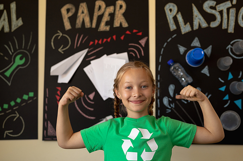 Portrait of a Caucasian schoolgirl with plaits wearing a green t shirt with a white recycling logo on it, standing in front of posters about recyclable materials, flexing her biceps and looking to camera smiling in an elementary school classroom