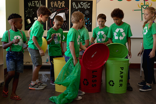 Side view of a diverse group of schoolchildren wearing green t shirts with a white recycling logo on them, holding color coded recycling bins and bags in an elementary school classroom