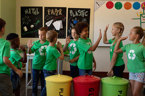 Side view of a diverse group of schoolchildren wearing green t shirts with a white recycling logo on them, standing beside colour coded recycling bins in an elementary school classroom, high fiving with each other and smiling