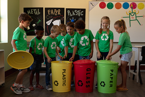 Side view of a diverse group of schoolchildren wearing green t shirts with a white recycling logo on them, holding colour coded recycling bins in an elementary school classroom