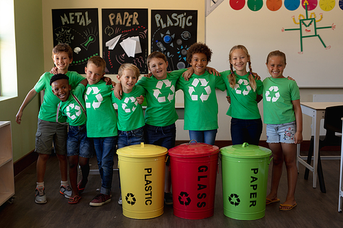 Portrait of a diverse group of schoolchildren wearing green t shirts with a white recycling logo on them, standing behind colour coded recycling bins in an elementary school classroom with arms around each other, smiling to camera