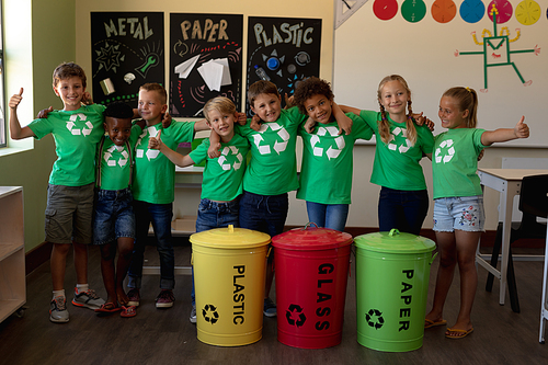 Portrait of a diverse group of schoolchildren wearing green t shirts with a white recycling logo on them, standing behind colour coded recycling bins in an elementary school classroom with arms around each other, smiling to camera with thumbs up