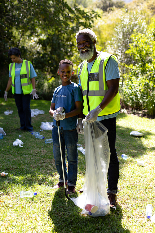 Senior African American man spending time outside with his family, embracing his grandson, looking at the camera and smiling, wearing blue volunteers t shirts, collecting garbage, holding a garbage bag, on a sunny day