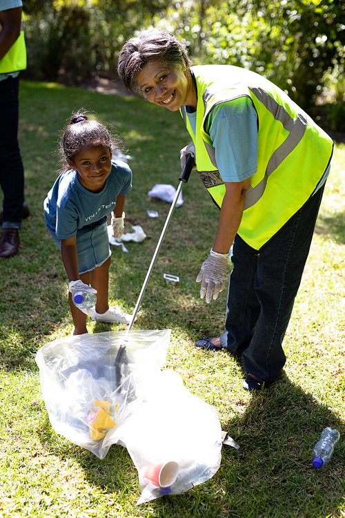 Senior mixed race woman spending time outside with her family, putting garbage into a bag with her granddaughter, wearing blue volunteers t shirts, looking at the camera and smiling, on a sunny day