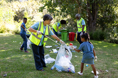 Senior mixed race woman spending time outside with her family, putting garbage into a bag with her granddaughter, wearing blue volunteers t shirts, on a sunny day