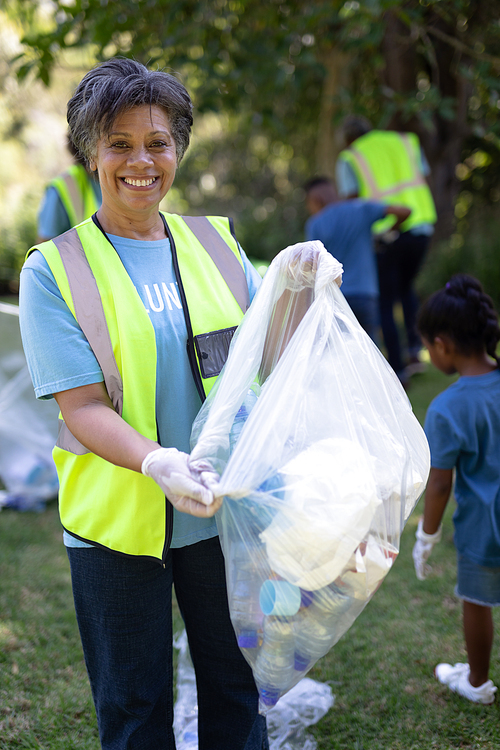 Portrait of a senior mixed race woman spending time outside with her family, presenting a garbage bag with garbage in it, looking at the camera and smiling, with her family in the background, all wearing blue volunteers t shirts, on a sunny day