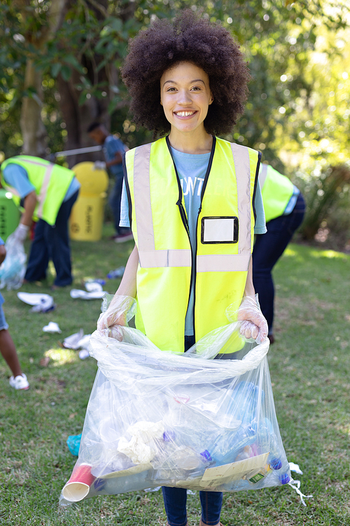 Portrait of a mixed race woman spending time outside with her family, presenting a garbage bag with garbage in it, looking at the camera and smiling, with her family in the background, all wearing blue volunteers t shirts, on a sunny day