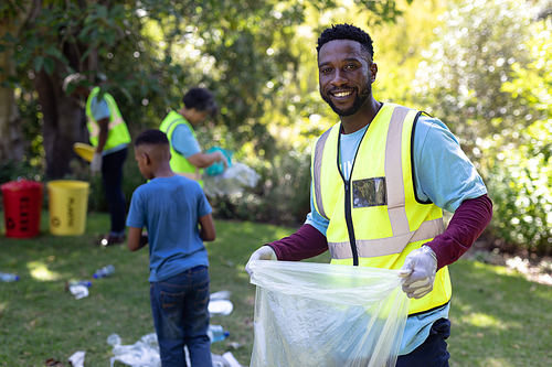 Portrait of an African American man spending time outside with his family, presenting a garbage bag with garbage in it, looking at the camera and smiling, with his family in the background, all wearing blue volunteers t shirts, on a sunny day