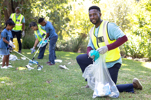 Portrait of an African American man spending time outside with his family, presenting a garbage bag with garbage in it, looking at the camera and smiling, with his family in the background, all wearing blue volunteers t shirts, on a sunny day