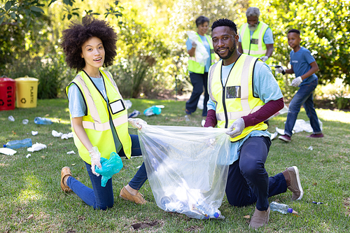 Portrait of a mixed race couple spending time outside with their family, presenting a garbage bag with garbage in it, looking at the camera and smiling, with their family in the background, all wearing blue volunteers t shirts, on a sunny day