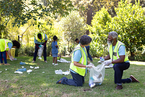 Senior mixed race couple spending time outside with their family, presenting a garbage bag with garbage in it, with their family in the background, all wearing blue volunteers t shirts, on a sunny day