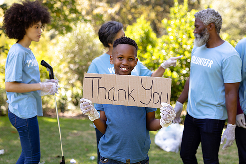 Mixed race boy spending time outside with his family, holding a thank you sign, looking at the camera and smiling, with his family in the background, all wearing blue volunteer t shirts, on a sunny day