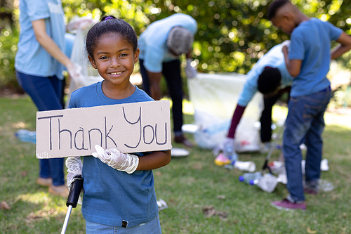 Mixed race girl spending time outside with her family, holding a thank you sign, looking at the camera and smiling, with her family in the background, all wearing blue volunteer t shirts, on a sunny day