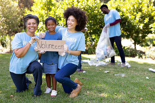 Mixed race girl spending time outside with her family, holding a volunteer sign with her mother and grandmother, looking at the camera and smiling, all wearing blue volunteer t shirts, on a sunny day
