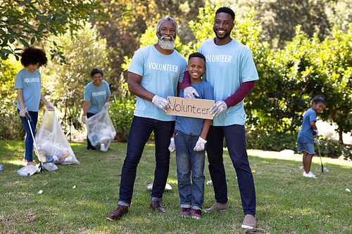 Mixed race boy spending time outside with his family, holding a volunteer sign with his father and grandfather, looking at the camera and smiling, all wearing blue volunteer t shirts, on a sunny day