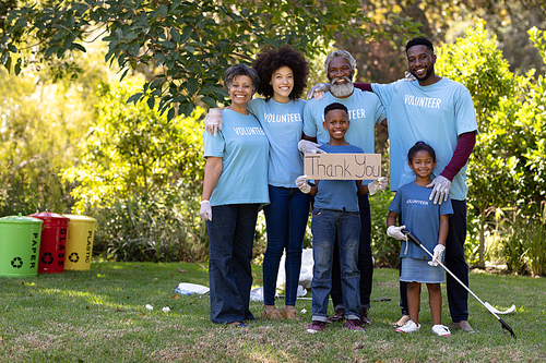 Multi-generation mixed race family spending time outside together, all wearing blue volunteer t shirts and protective gloves, collecting garbage, standing in a row, embracing each other, holding a volunteer sign, on a sunny day