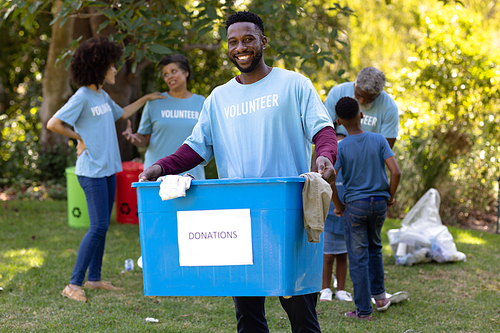 Portrait of an African American man spending time outside with his family, presenting a donations box with clothes in it, looking at the camera and smiling, with his family in the background, all wearing blue volunteers t shirts, on a sunny day