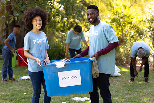 Mixed race couple spending time outside with their family, presenting a donations box with clothes in it, looking at the camera and smiling, with their family in the background, all wearing blue volunteers t shirts, on a sunny day