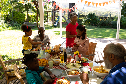 Side view of a multi-ethnic, multi-generation family serving food, sitting down at a table and talking during a meal together outside on a patio in the sun