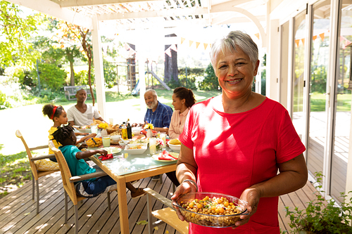 A mixed race senior woman wearing a red dress standing on a patio in the sun holding a dish of bean salad and smiling to camera, multi-generation family sitting down at a table for a meal outside