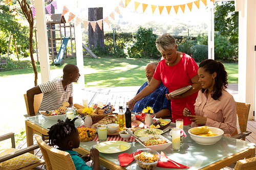 Side view of a multi-ethnic, multi-generation family serving food and sitting down at a table for a meal outside on a patio in the sun, the grandmother standing and putting plates on the table