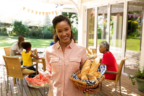 a mixed race woman standing on a patio in the sun holding a bread basket and a plate of sliced  and smiling to camera, multi-generation family sitting down at a table for a meal outside