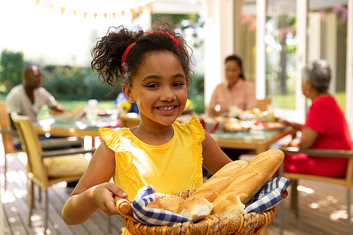A mixed race girl wearing a yellow dress standing on a patio in the sun holding a bread basket and smiling to camera, multi-generation family sitting down at a table for a meal together outside