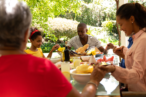 Over the shoulder view of a multi-ethnic, multi-generation family sitting down at a table holding hands with eyes closed and saying a prayer before having a meal together outside on a patio in the sun