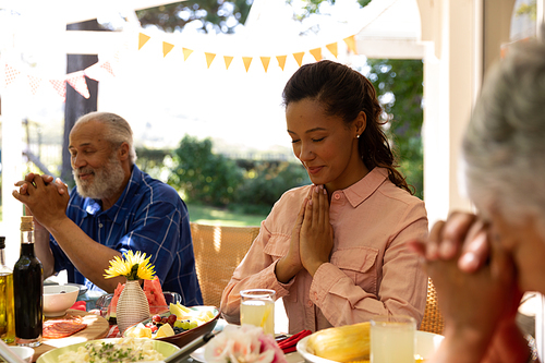 A multi-ethnic, multi-generation family sitting down at a table with eyes closed and hands in prayer, saying grace before having a meal outside on a patio in the sun, in the foreground a senior woman