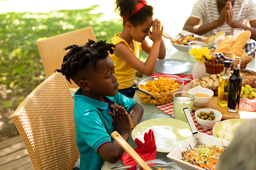 A young African American brother and sister sitting at a table with eyes closed and hands in prayer, saying grace before having a meal, multi-generation family outside on a patio