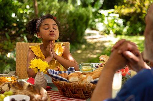 An African American girl sitting at a table with eyes closed and hands in prayer, saying grace before having a meal with her family outside on a patio in the sun, her grandfather in the foreground
