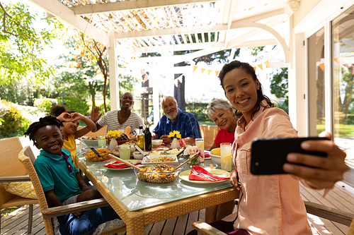 A multi-ethnic, multi-generation family sitting at a table for a meal together outside on a patio in the sun, the mixed race mother using a smartphone to take a selfie with them all smiling