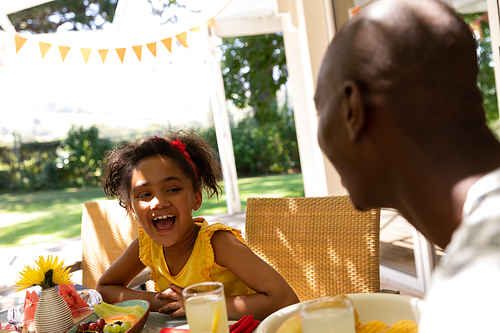 Side view of an African American man and his young daughter smiling and laughing, sitting at a table for a family meal outside on a patio in the sun