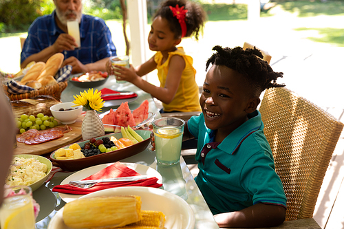 Side view of a smiling young African American boy and his sister sitting at a table with their grandfather for a family meal together outside on a patio in the sun