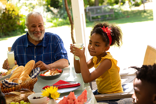 Side view of a young African American girl holding a glass sitting at a table with her young brother and smiling grandfather for a family meal outside on a patio in the sun