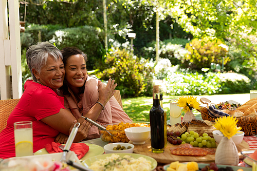 Front view of a mixed race woman and her senior mother embracing and smiling sitting at a table for a family meal outside on a patio in the sun