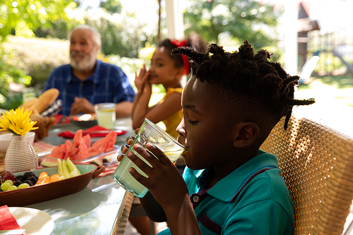A young African American boy holding a glass with two hands and drinking, sitting at a table with his multi-ethnic, multi-generation family for a meal together outside on a patio in the sun
