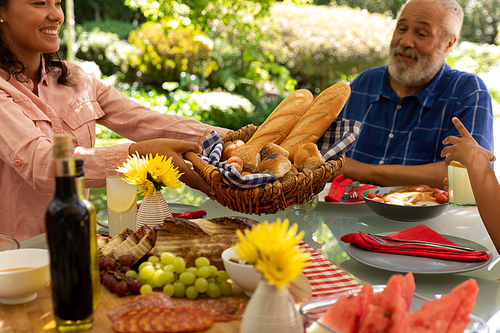 Side view of a mixed race woman sitting at a dinner table offering a basket of bread, her senior father sitting beside her smiling, during a family meal outside on a patio in the sun