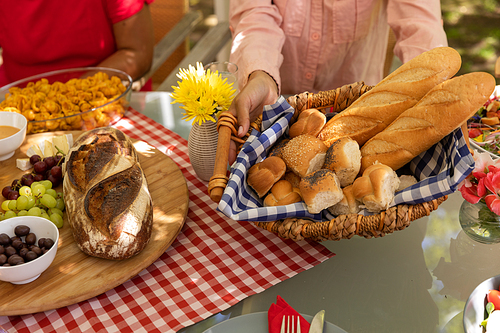 High angle mid section of a mixed race woman holding a basket of bread sitting at a table for a family meal outside on a patio in the sun
