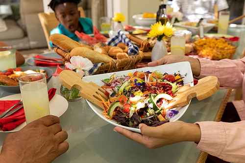 Side view of the hands of a mixed race woman passing a bowl of salad, sitting at a table during a family meal outside on a patio in the sun, her young African American son sitting in the background