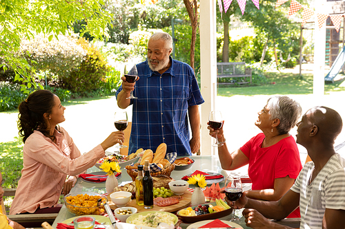 A mixed race senior man standing, raising a glass of red wine and making a toast with his multi-ethnic adult family sitting at the table and raising their glasses during a meal outside on a patio