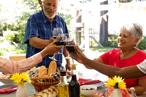 A mixed race senior man standing, holding a glass of red wine and making a toast with his multi-ethnic adult family, sitting at the table and raising their glasses during a meal outside on a patio