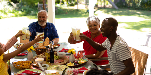 Side view of a multi-ethnic, multi-generation family sitting at a table for a meal together outside on a patio in the sun, raising glasses of lemonade and making a toast