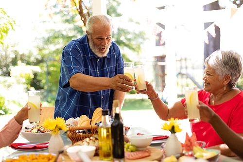A mixed race senior man raising a glass of lemonade and making a toast with his multi-ethnic adult family, sitting at the table and raising their glasses during a meal together outside on a patio