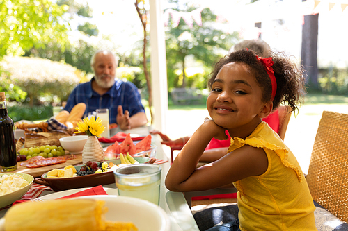 A young mixed race girl wearing a yellow dress turning and smiling to camera sitting at a table during a family meal outside on a patio in the sun, her grandparenst sitting in the background