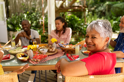 Side view of a senior mixed race woman turning and smiling to camera, sitting at a table during a family meal outside on a patio in the sun, her adult children in the background