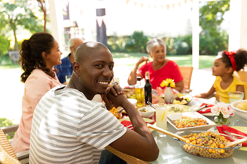 Side view of an African American man turning to camera and smiling, sitting sitting at a table during a meal with his multi-ethnic, multi-generation family outside on a patio in the sun