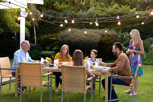 Side view of a Caucasian multi-generation family sitting at a table for a meal together outside in the garden, the mother standing beside her husband