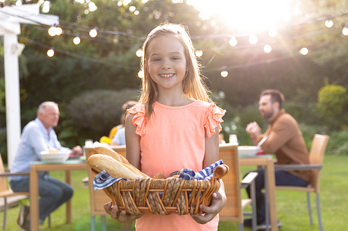 A Caucasian girl with long blonde hair standing in the garden holding a basket of bread and smiling to camera, in the background her multi-generation family sitting at a table for a meal outside