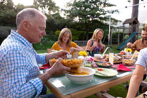 Side view close up of a senior Caucasian man sitting at a table serving food during a multi-generation family meal in the garden, his relatives serving food and eating in the background
